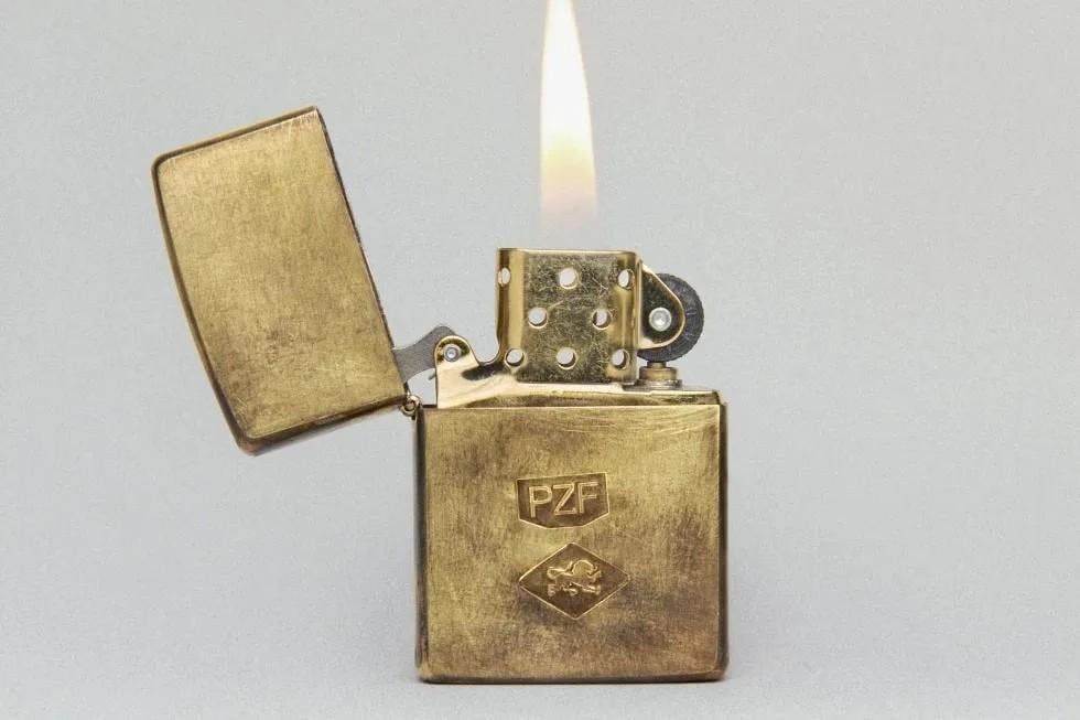 A close-up photo of a lit gold Zippo lighter, with the letters PZF engraved on it.

