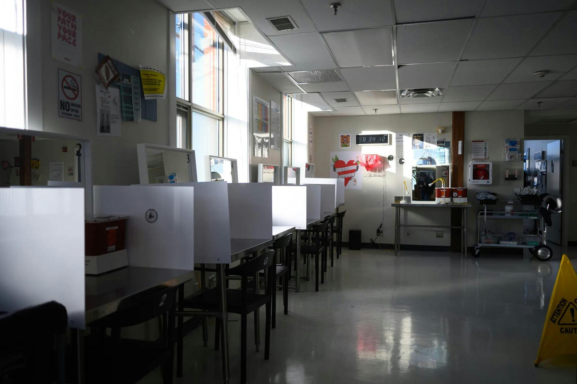 Image of sunlight coming through the windows into a dark room furnished with chairs, mirrors and tables separated by white screens and decorated with posters on the walls. 
