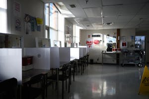 Image of sunlight coming through the windows into a dark room furnished with chairs, mirrors and tables separated by white screens and decorated with posters on the walls. 
