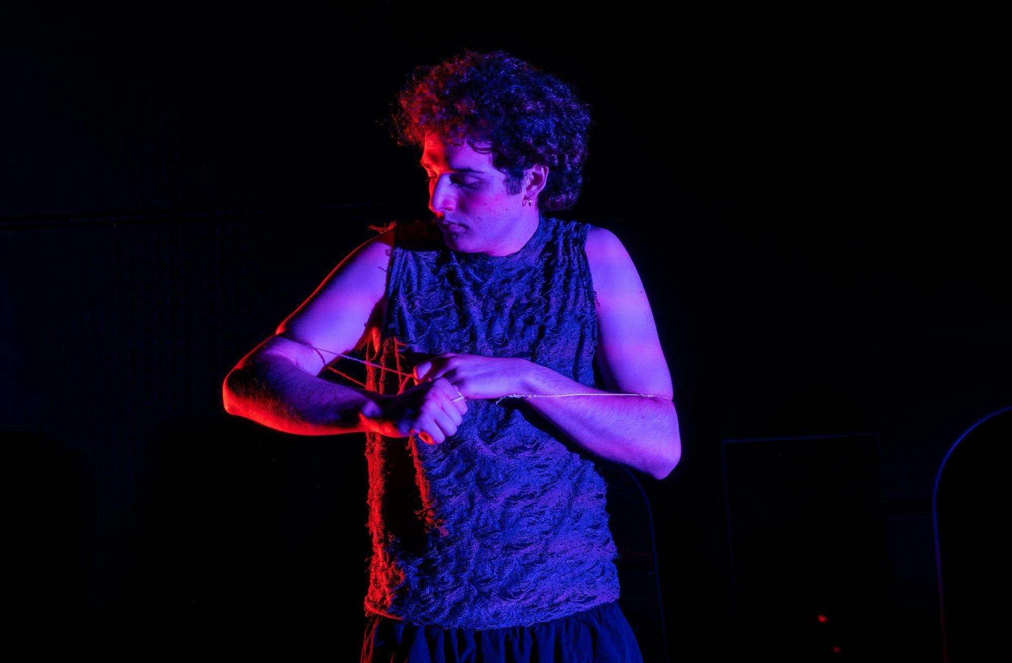 Photo of a curly-haired man looking to the left bathed in a violet light with a string tied loosely around his chest and arms.