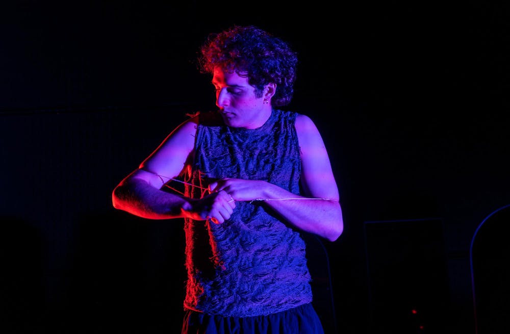 Photo of a curly-haired man looking to the left bathed in a violet light with a string tied loosely around his chest and arms.