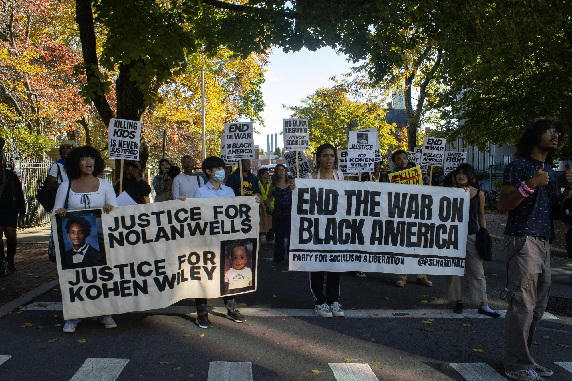 Photo of a crowd of people walking through the street holding signs. The three people in front hold large white banners that read “JUSTICE FOR NOLAN WELLS”, “JUSTICE FOR KOHEN WILEY” and “END THE WAR ON BLACK AMERICA” in black letters. 