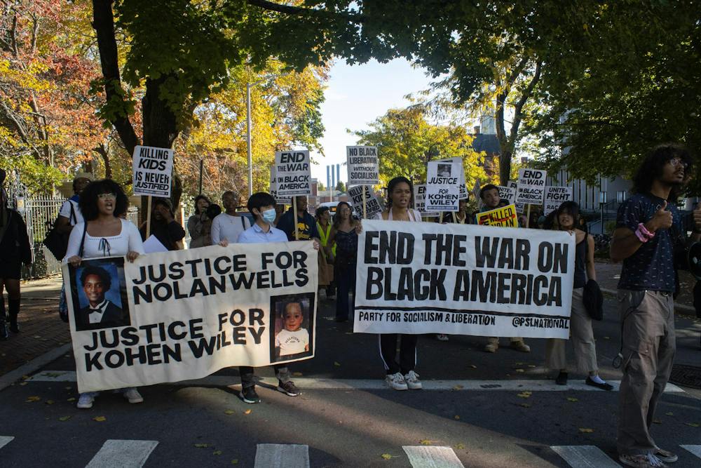 Photo of a crowd of people walking through the street holding signs. The three people in front hold large white banners that read “JUSTICE FOR NOLAN WELLS”, “JUSTICE FOR KOHEN WILEY” and “END THE WAR ON BLACK AMERICA” in black letters. 