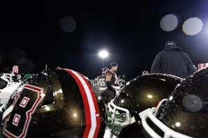 A photograph with three Brown football helmets in the foreground, and a single Brown player framed by the helmets standing farther away. There is a floodlight in the background.