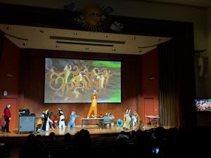 A photo of computer science teaching assistants dressed as Lion King-themed characters bowing around a table. One TA in a costume holds a plush toy in the air, standing on the table. A scene of animals from The Lion King plays in the background. 