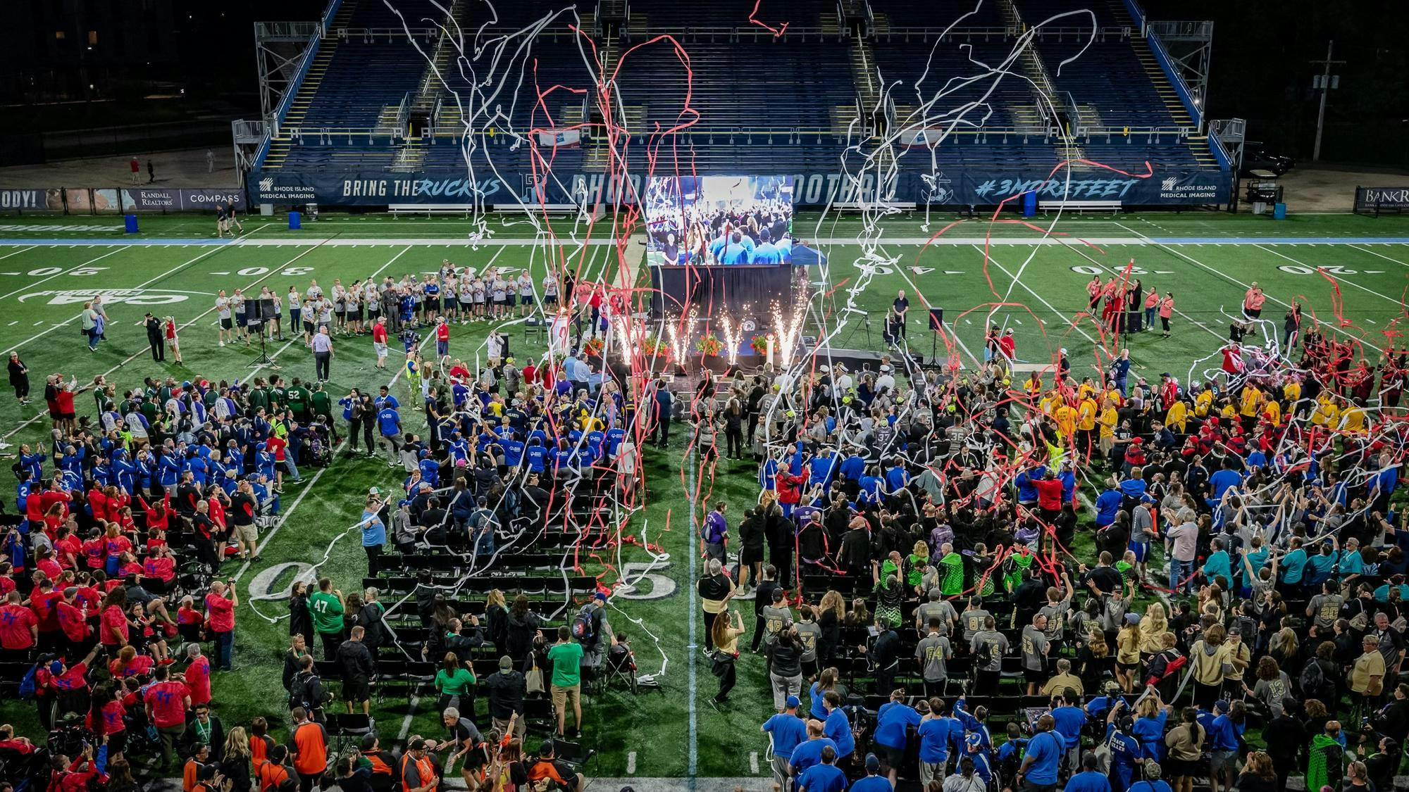Crowds of athletes stand in a stadium as multicolored confetti and fireworks explode into the air.