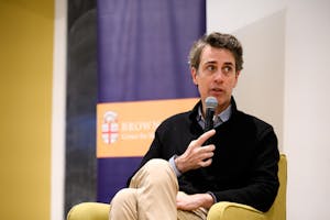 Photo of Ben Moser in a blue collared shirt, black quarter zip and khakis, holding a microphone while sitting in a yellow armchair.