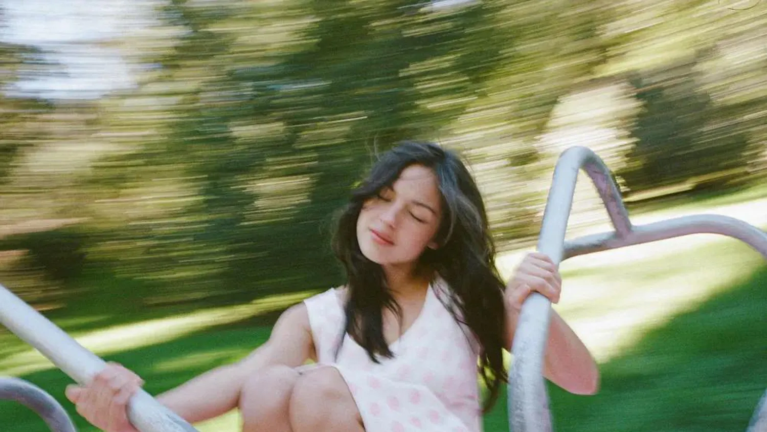 A promotional photograph of Olivia Rodrigo spinning on a playground merry-go-round.