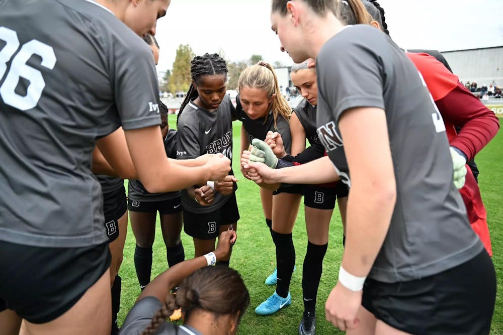 Around ten Brown women's soccer players are gathered around in a circle with their fists outstretched as they get ready to break and return to play. 