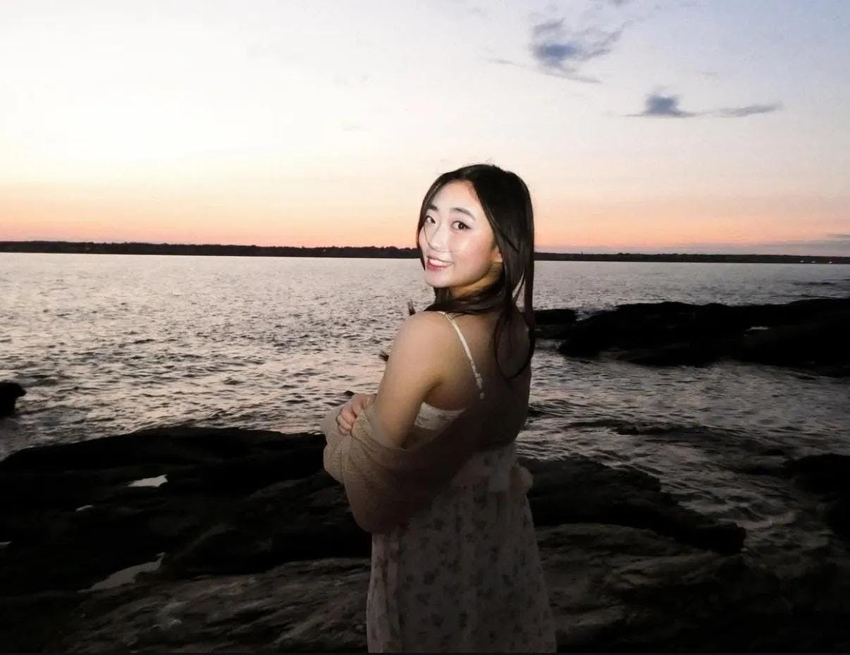 Photo of a girl wearing a white floral sundress and a brown cardigan facing the sunset and sea with her head turned towards the left, looking at the camera.