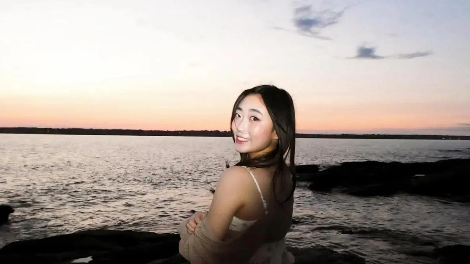 Photo of a girl wearing a white floral sundress and a brown cardigan facing the sunset and sea with her head turned towards the left, looking at the camera.