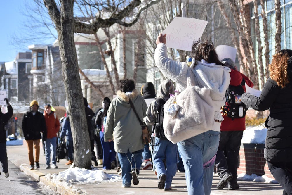 A group of student protesters walking down the sidewalk. One protester holds up a sign that states “Pueblo / abre los / ojos!!!”