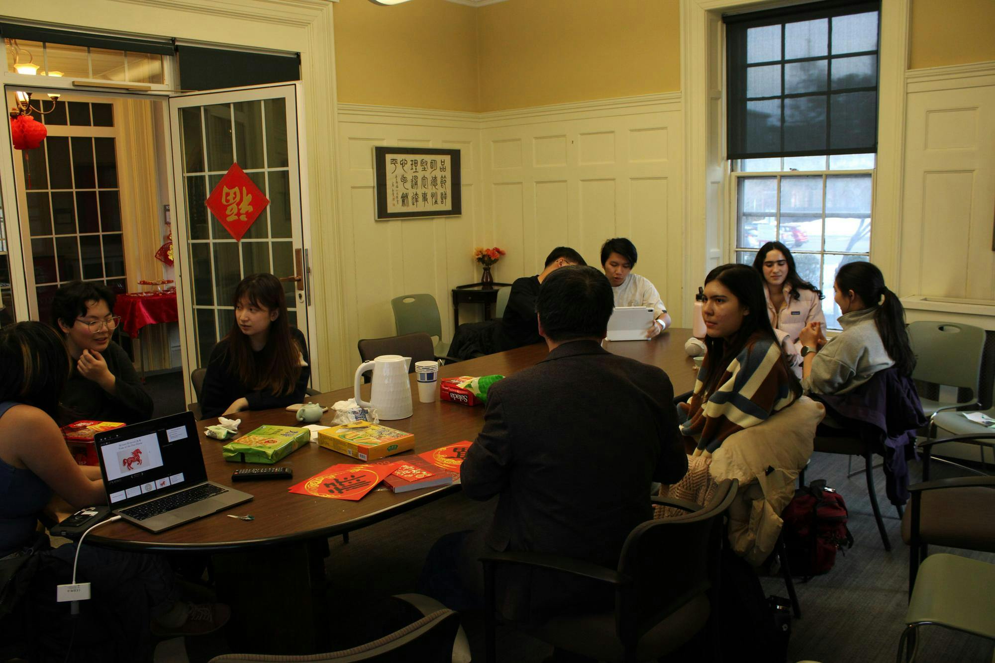 Photo of students around a long, ovular table speaking to each other with snacks and a white kettle on the table.

