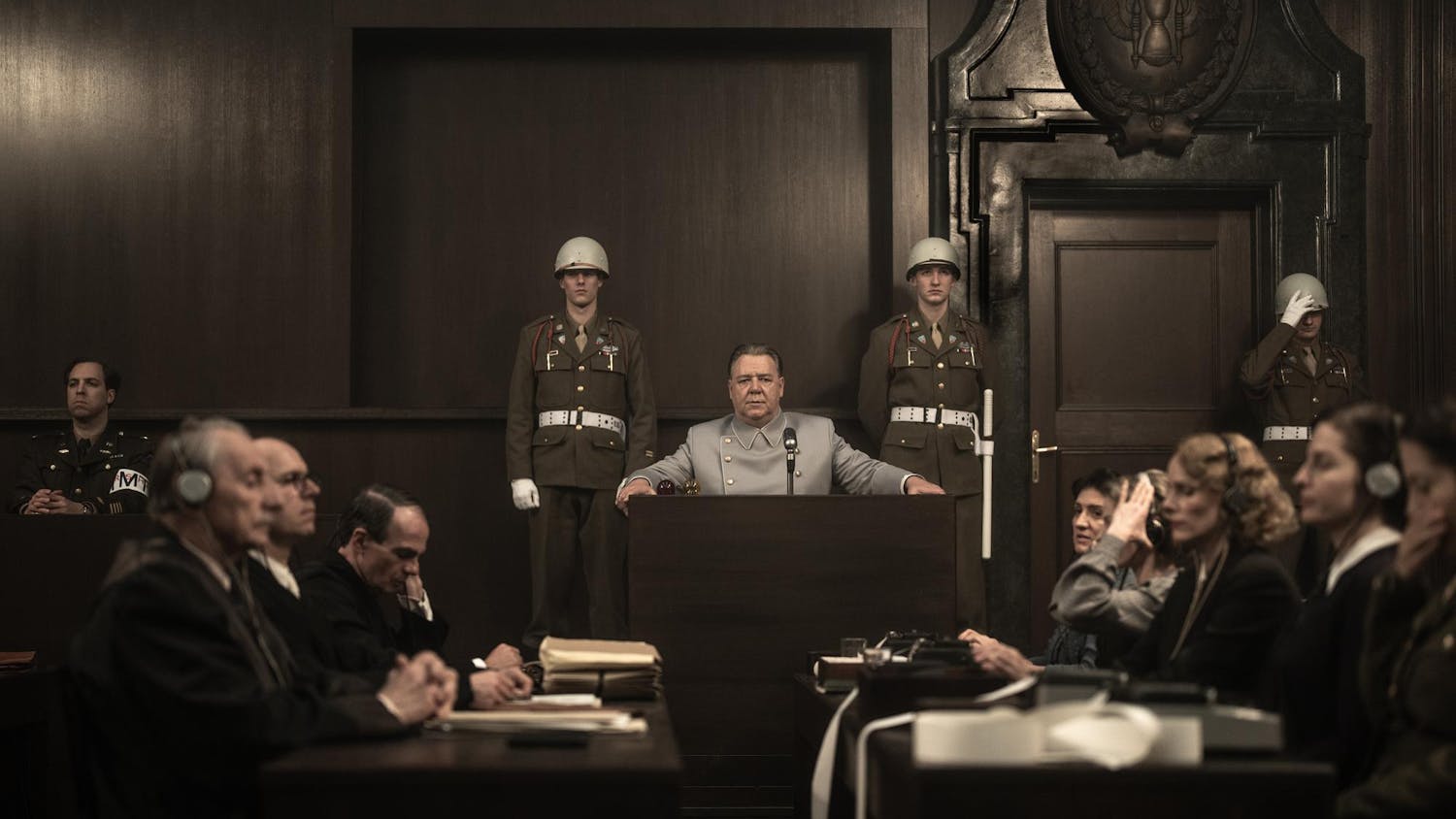 An older man sits at a podium in a courtroom. In front of the podium is a table of people transcribing and writing. On both sides of the podium are military officers.