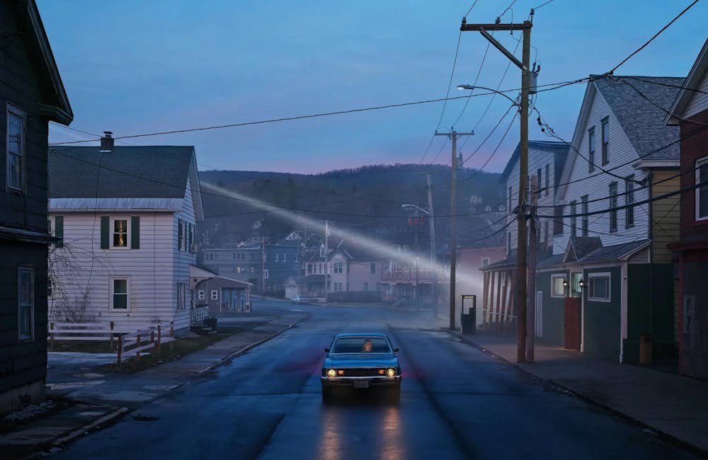 An artistic promotional photo of Phoebe Bridgers in a car at dusk driving down a long street alone.