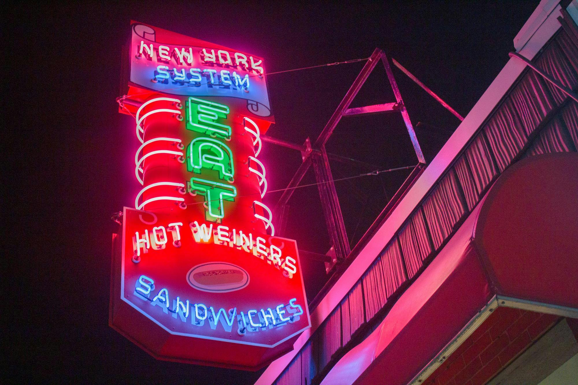 A neon sign, lit up at night, reads "New York System, Eat Hot Wieners, Sandwiches."