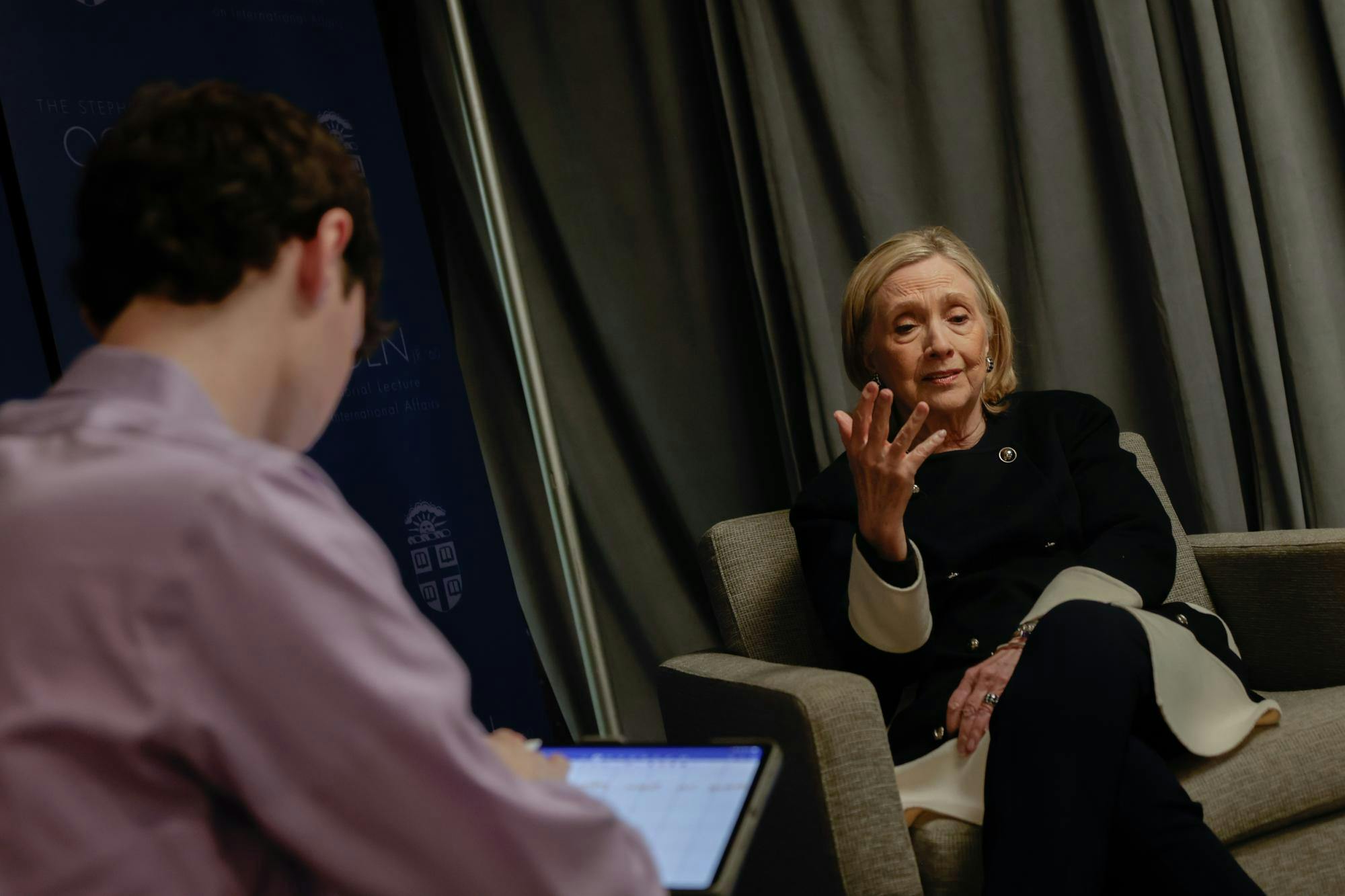 Photo of Hillary Clinton, viewed over the shoulder of someone in a lavender shirt, sitting in a grey chair raising one hand slightly as she speaks.