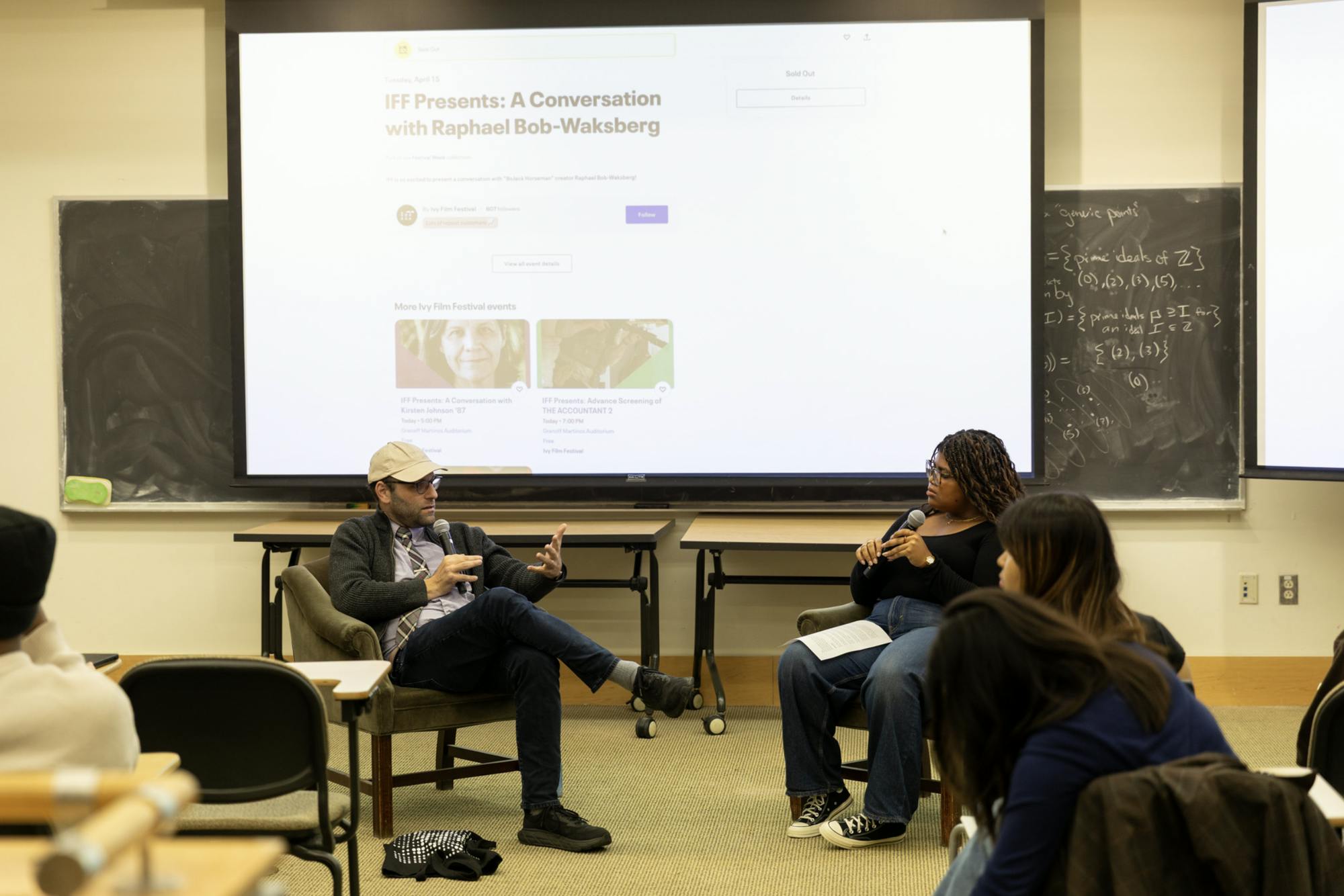 Raphael Bob-Waksberg, wearing a khaki baseball hat and jeans, sits in a chair across from moderator Gabriella Wrighten. They are in conversation in front of a brightly lit presentation. 