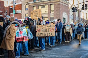 A line of protestors holding various signs. Many protestors wear surgical masks. 