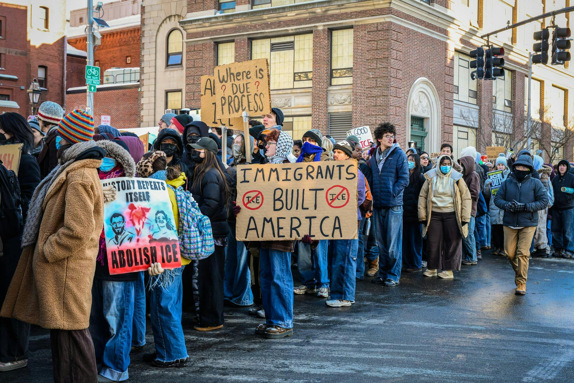 A line of protestors holding various signs. Many protestors wear surgical masks. 