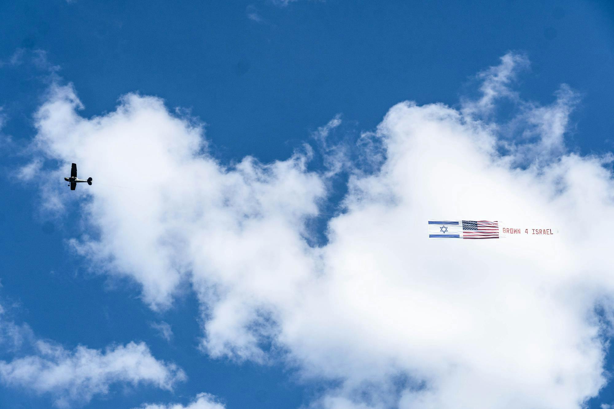 A photo shows a plane in a blue cloudy sky towing the American and Israeli flags, followed by red lettering reading “Brown 4 Israel.”