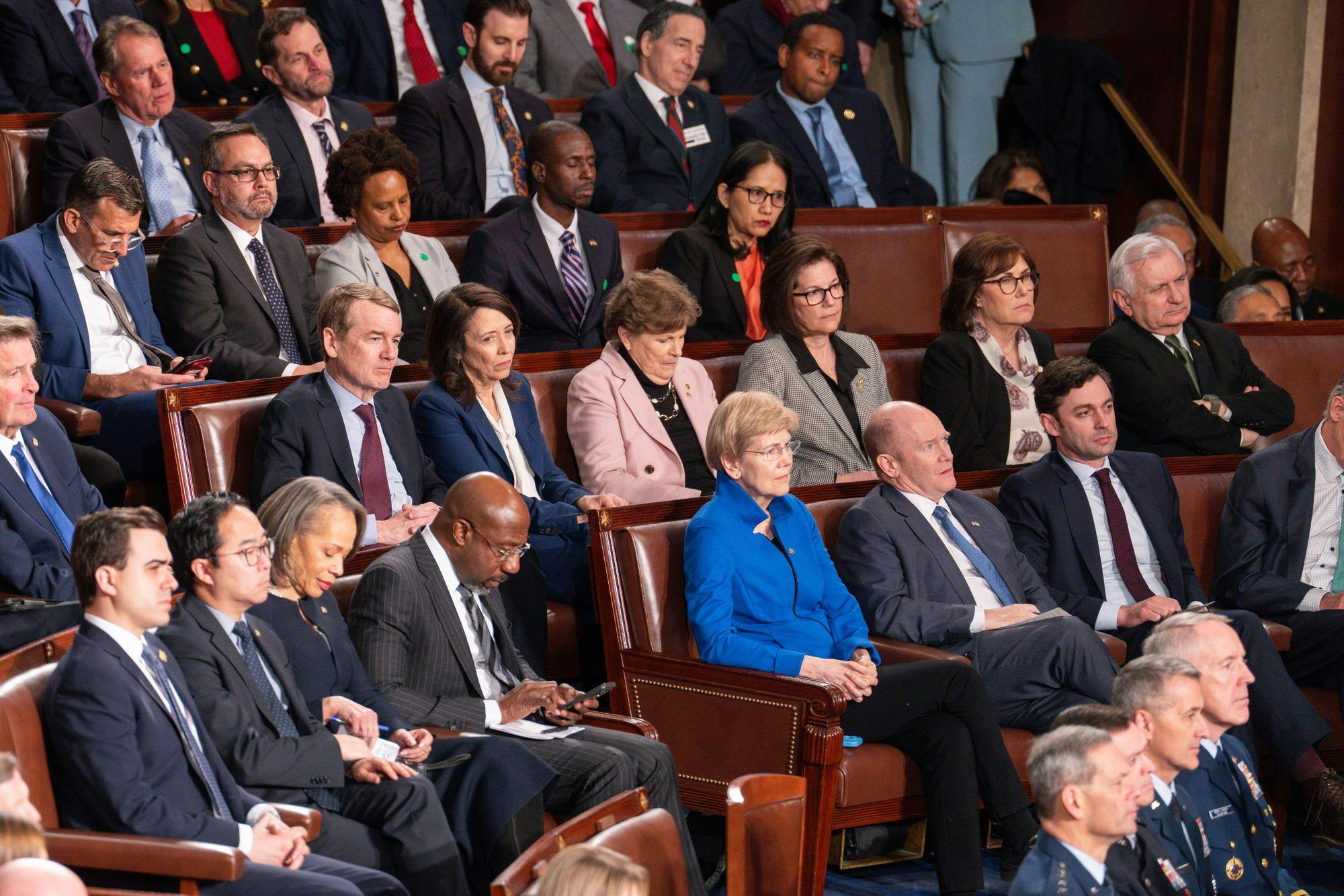 People in official attire seated in multiple rows of brown leather chairs in the House Chamber with an attentive expression on their faces. 