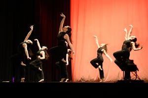 Multiple dancers dressed in all black posing with one foot up to the thigh and bending back with one arm raised.