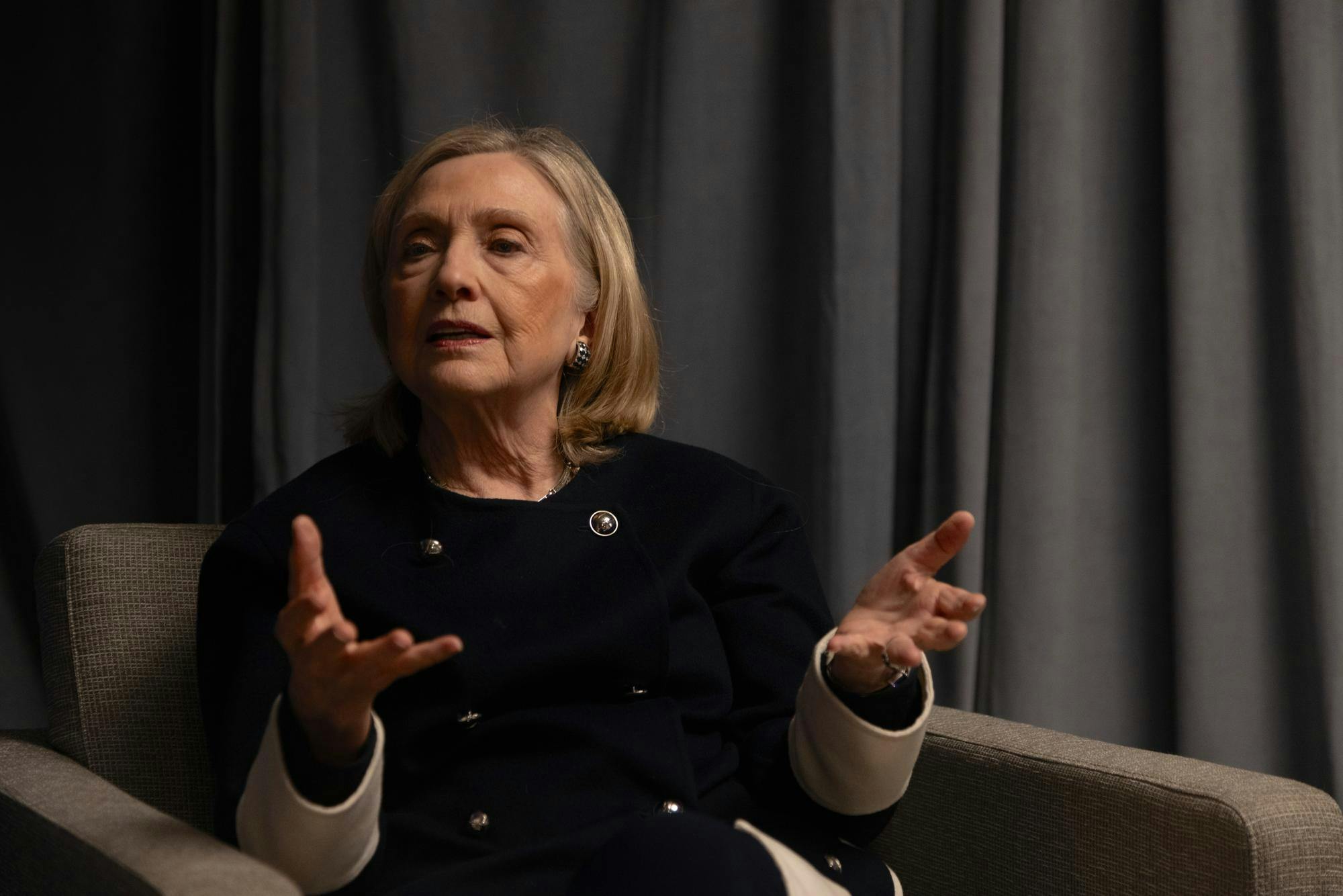 Photo of Hillary Clinton sitting in a grey chair and gesticulating with both hands