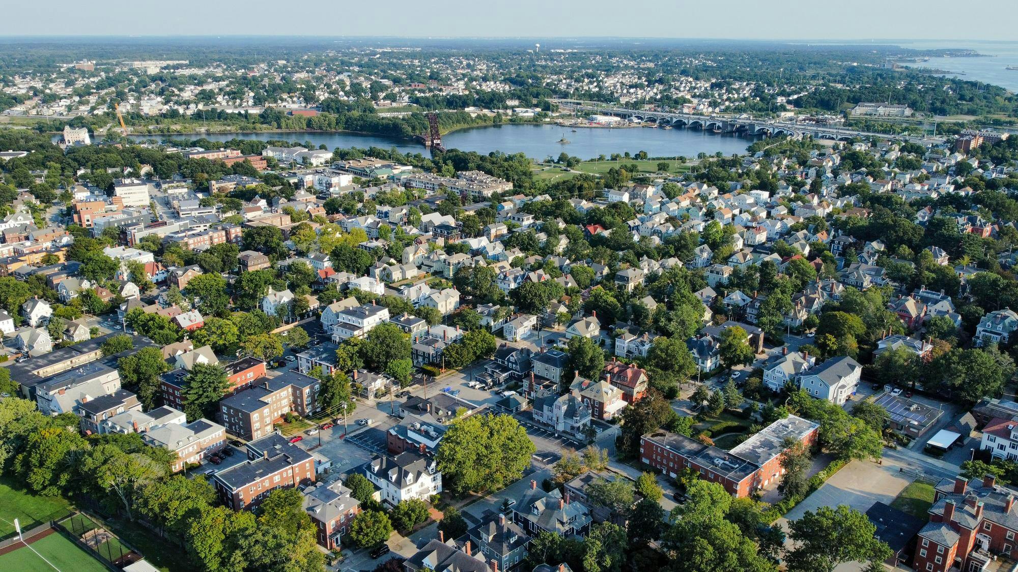 The picture depicts Rhode Island from above. The Washington Bridge looms far away. White and red houses speckle the landscape. The neighborhood is interspersed with trees and greenery. 