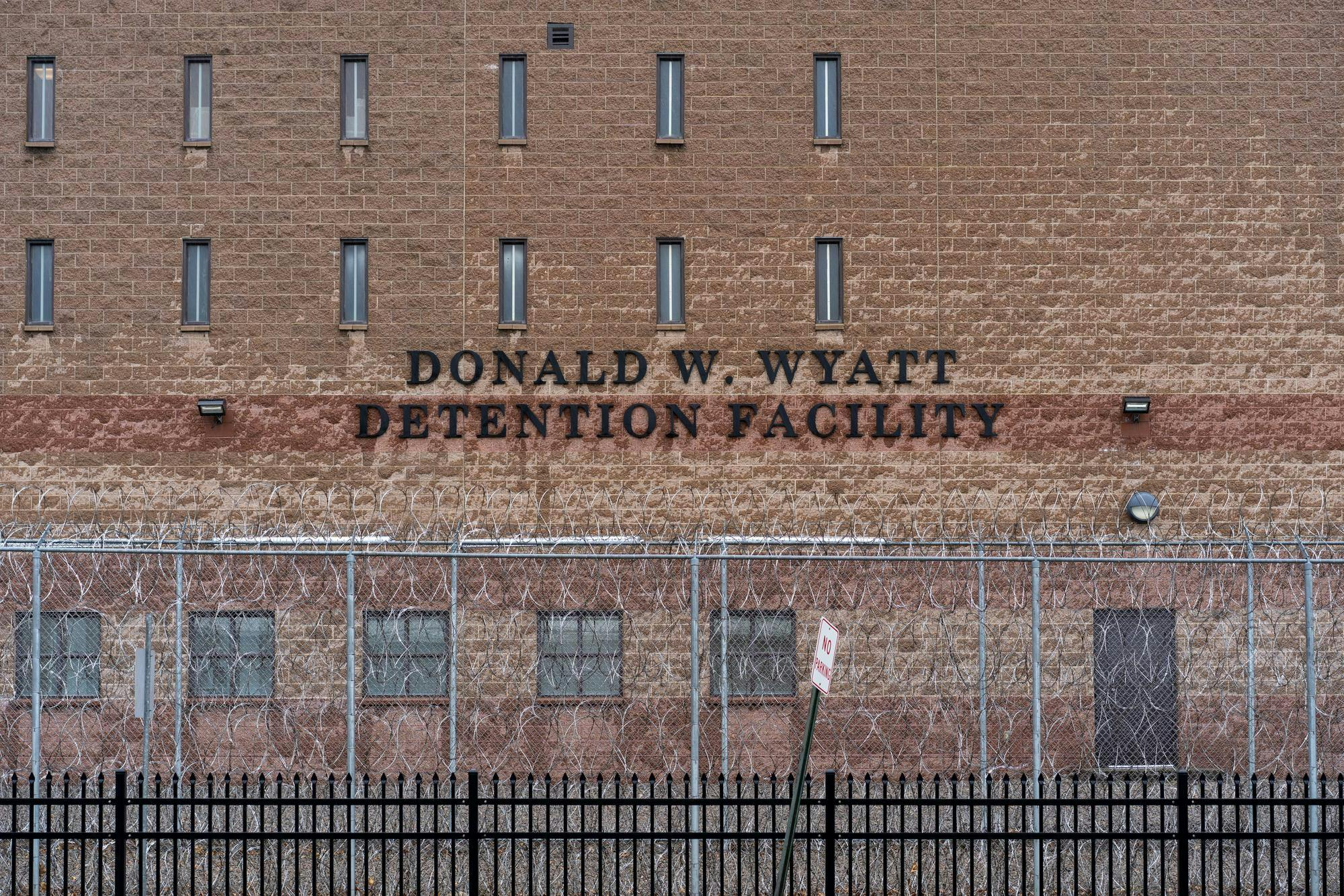 An image of a brick wall with "Donald W. Wyatt Detention Facility" written, a fence running along the front.
