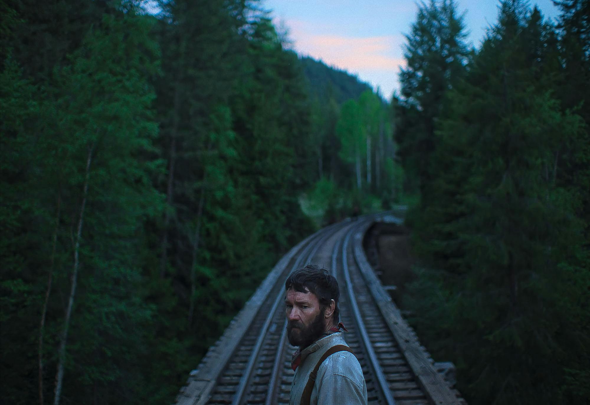 A photo shows Joel Edgerton as Robert Grainier, a bearded logger framed in the center of train tracks surrounded by trees. 