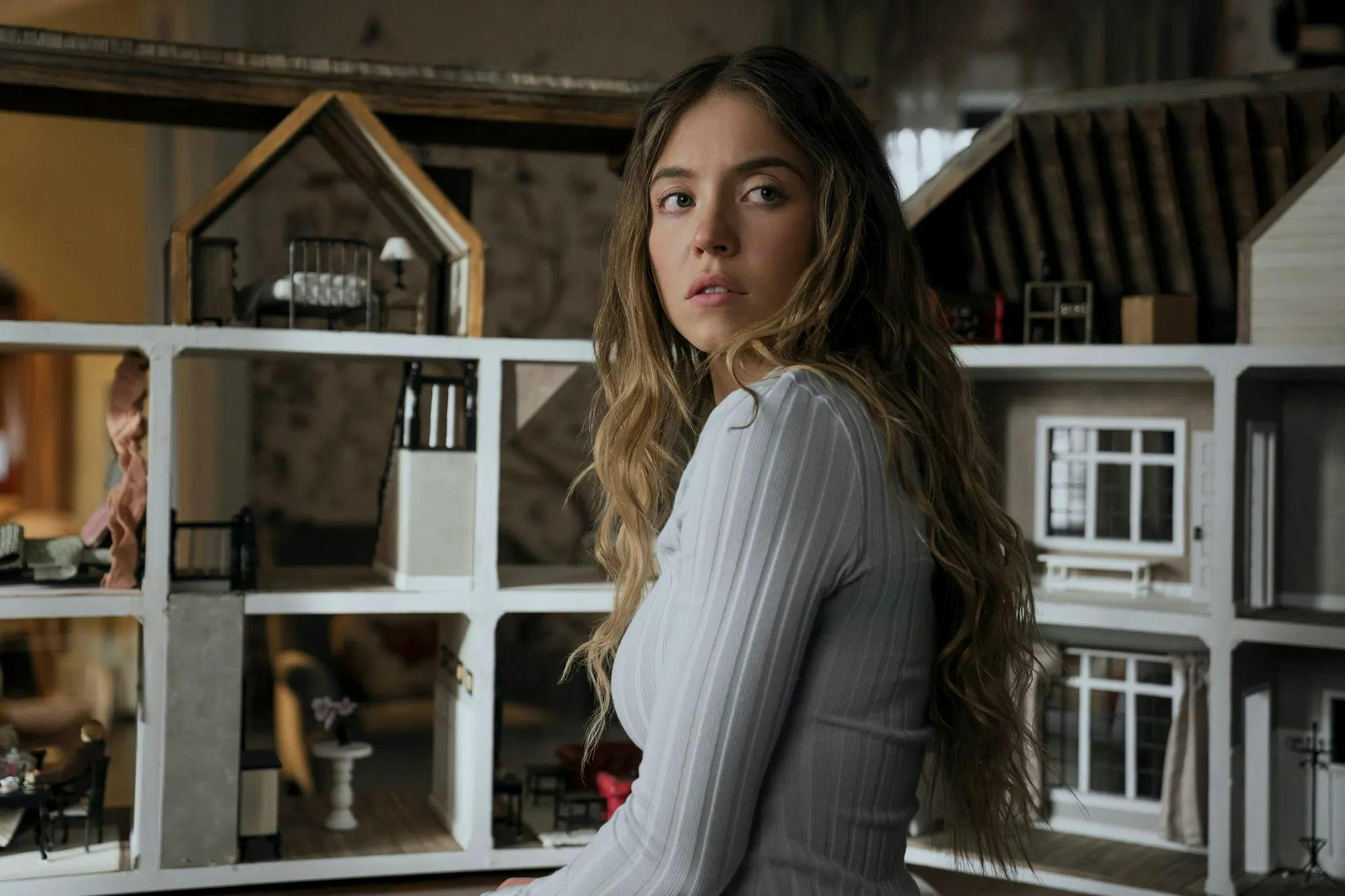 In a still from the movie, Sydney Sweeney, a blonde actress, wearing a white long-sleeved shirt, is pictured in front of a miniature model of a house. 
