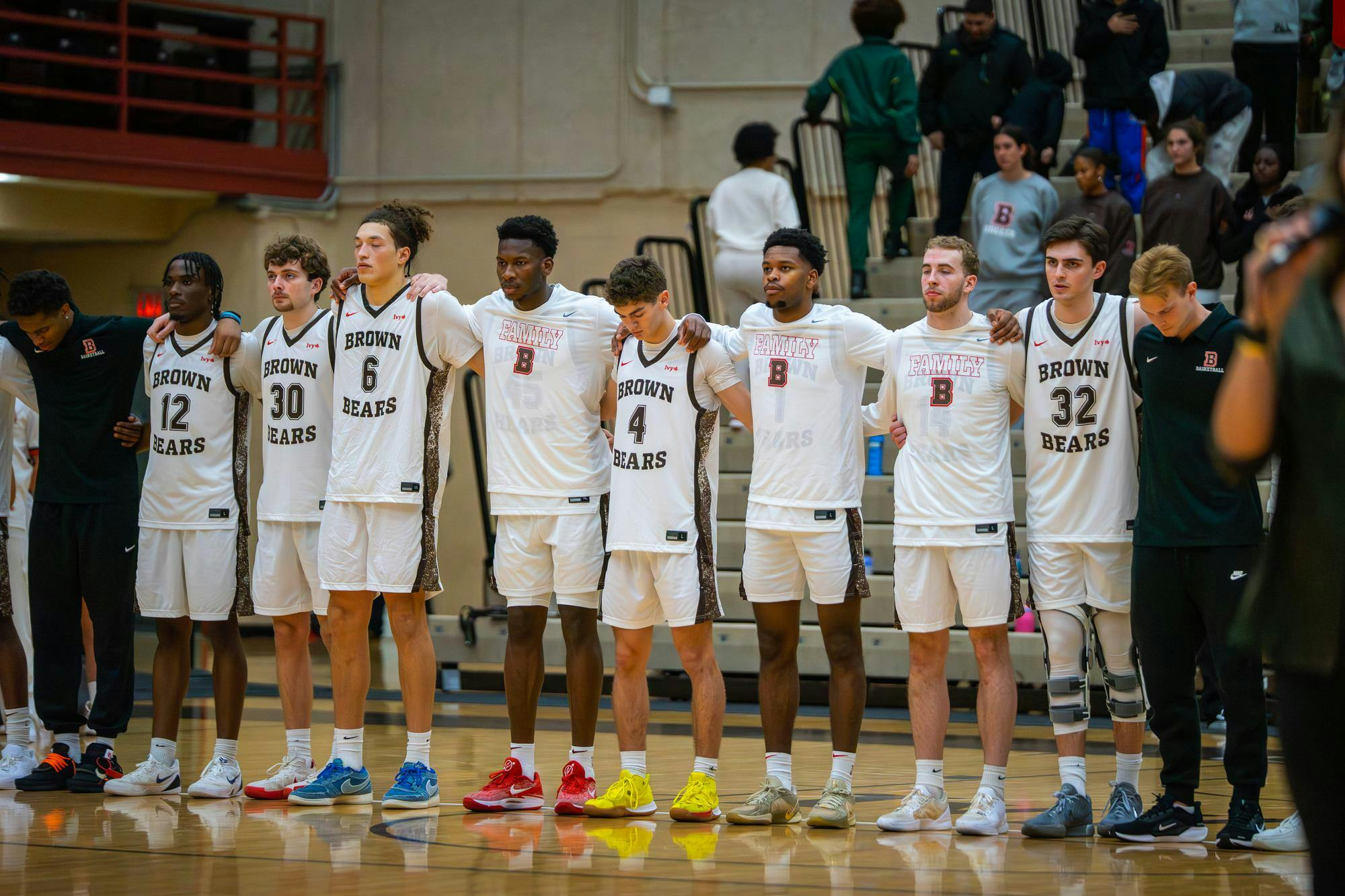 Photo of Brown University Men’s Basketball Team standing in line, arms interlocked, facing towards the left of the camera.