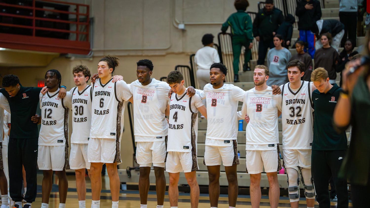 Photo of Brown University Men’s Basketball Team standing in line, arms interlocked, facing towards the left of the camera.