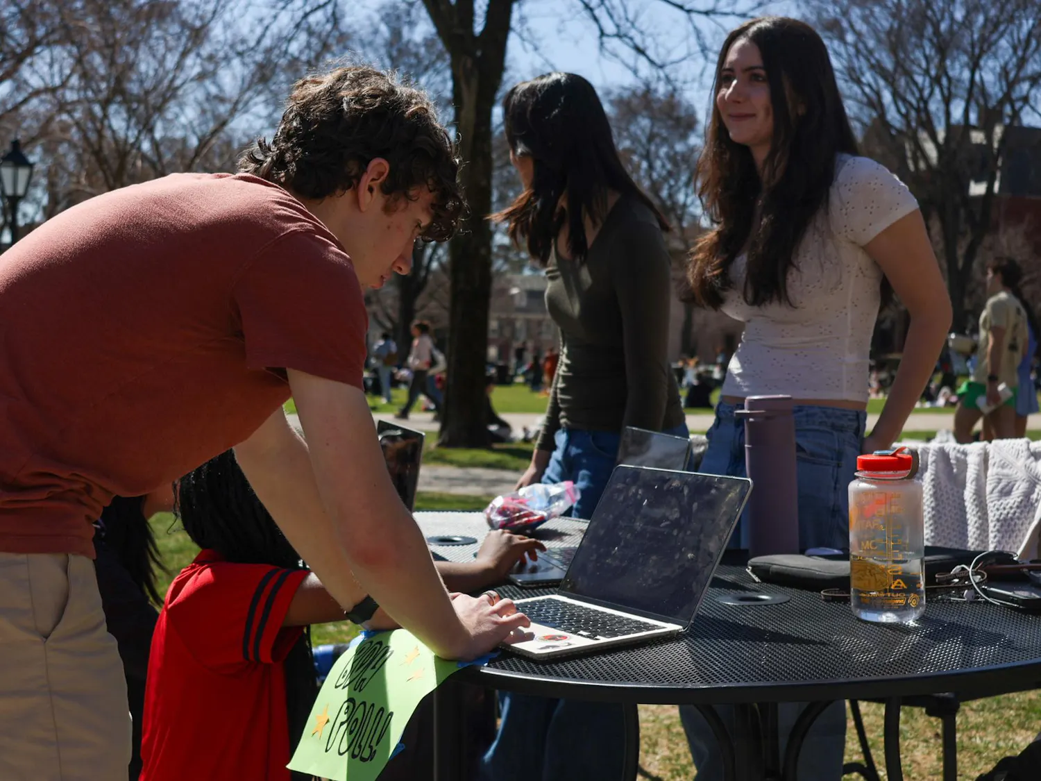A male undergraduate with curly brown hair is filling out The Herald's spring poll at a polling station on the Main Green. The sun is shining, and two Herald staffers, one wearing a white blouse, the other a dark green shirt, are standing behind the polling station.