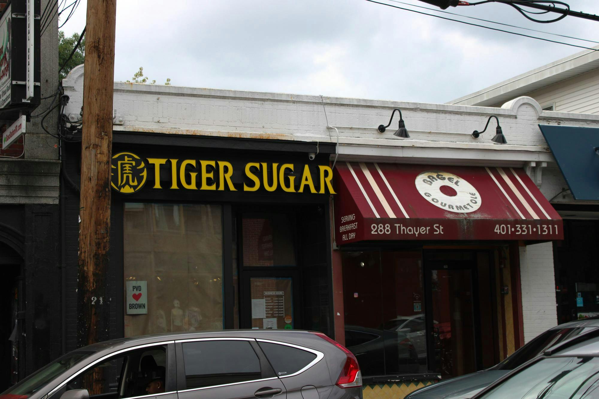Photo of two storefronts. The one on the left has big yellow words “Tiger Sugar” in all capital letters. The windows in the front are covered in brown paper with a PVD heart Brown sign. The store on the right has a red awning with white stripes and a white bagel with the words “Bagel Gourmet” in all capital letters. The store’s location and phone number are written below on the awning.