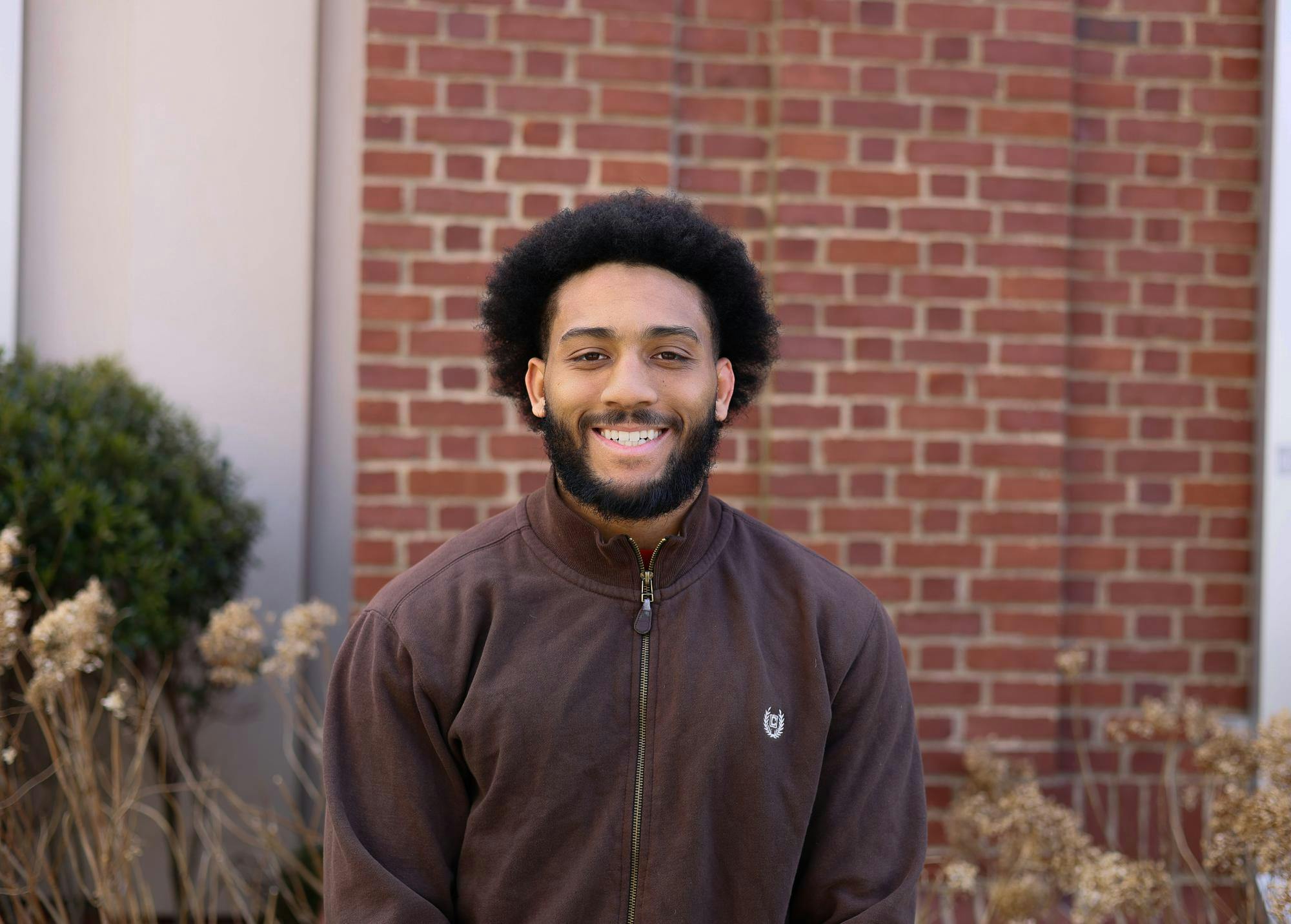 A photo of a man sitting outside a brick building wearing a brown sweater, smiling at the camera.