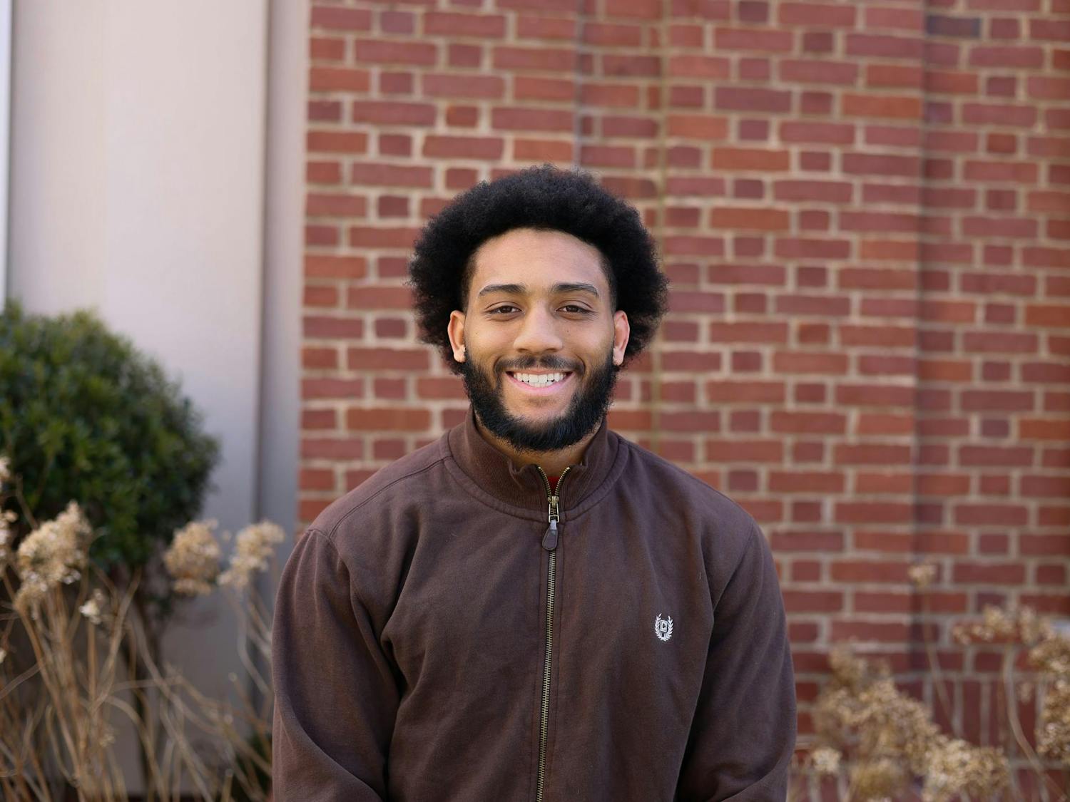 A photo of a man sitting outside a brick building wearing a brown sweater, smiling at the camera.