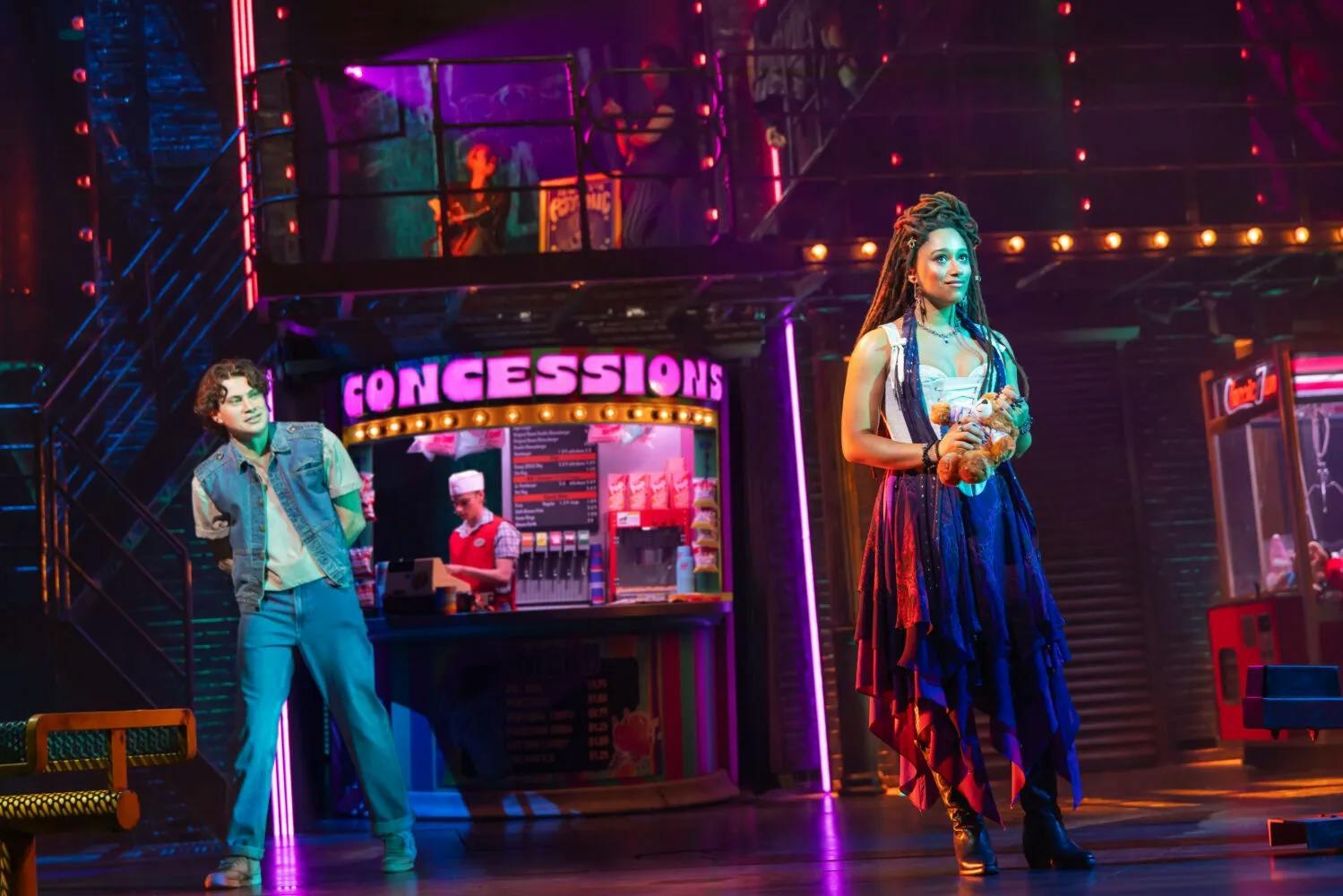 Photo of a performance of “The Lost Boys” on Broadway showing a woman in a purple dress and white corset holding a teddy bear looking toward the left. A man wearing a denim jacket and jeans is standing behind her, peering at her.