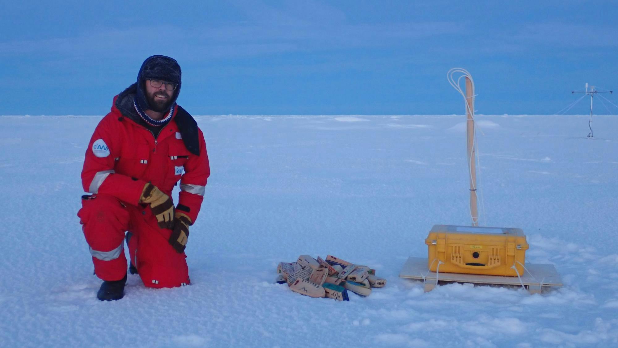 Photo of a male researcher wearing a red snowsuit and a winter hat smiling and kneeling on a large, snowy expanse with research equipment next to him.