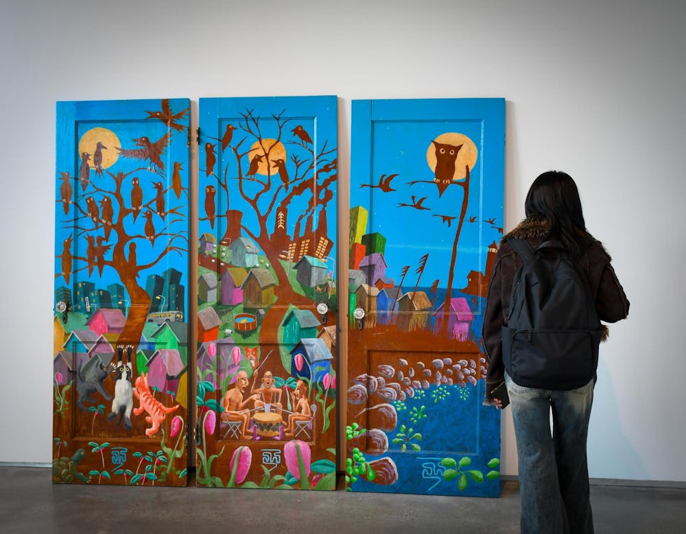 Photo of a girl wearing a backpack facing a wall. Leaning on the wall are three doors that have had a vibrant landscape of trees and houses painted on them.