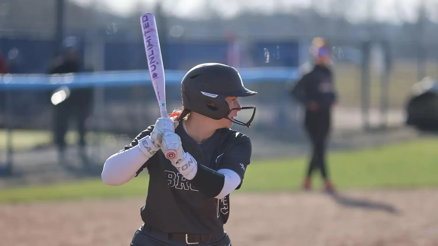 A Brown softball player in a black uniform, black helmet, and white sleeves holds up a white bat, prepared to swing.