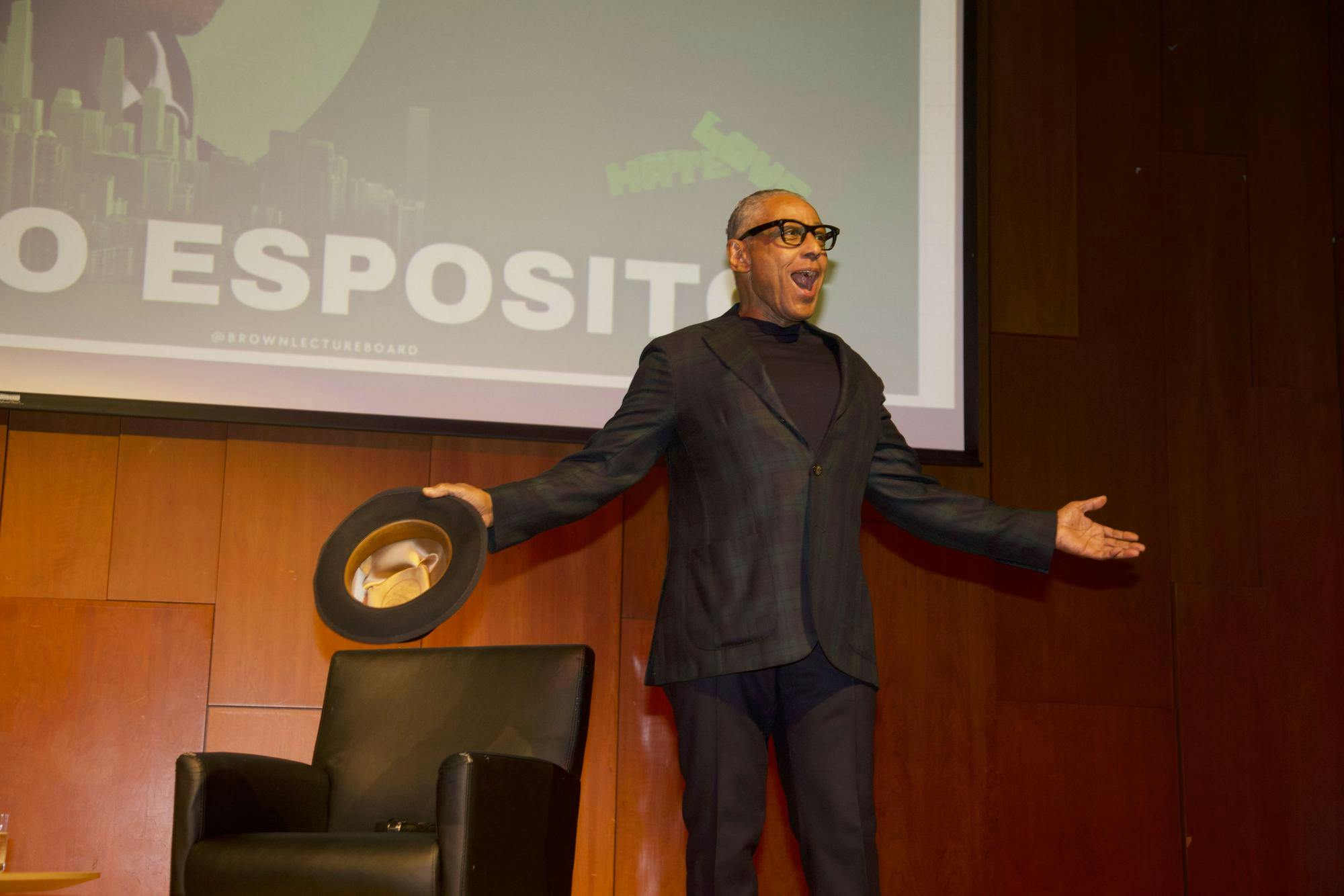 A photo of man dressed in all black opening his arms with a hat in his right hand. 