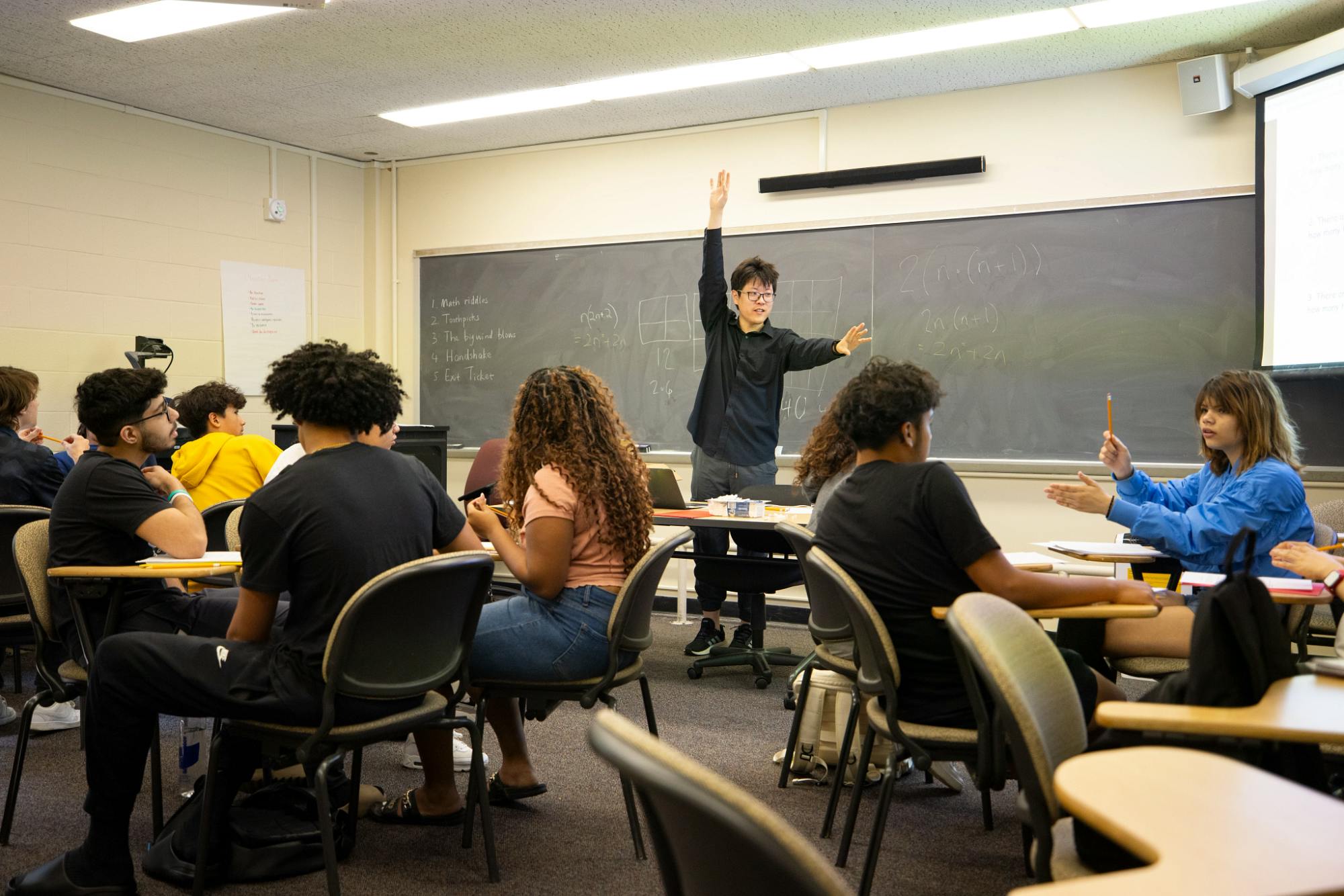 A black shirt-clad student stands at the front of his classroom directing his classmates. Behind him, a blackboard with various notes and diagrams drawn on it. 