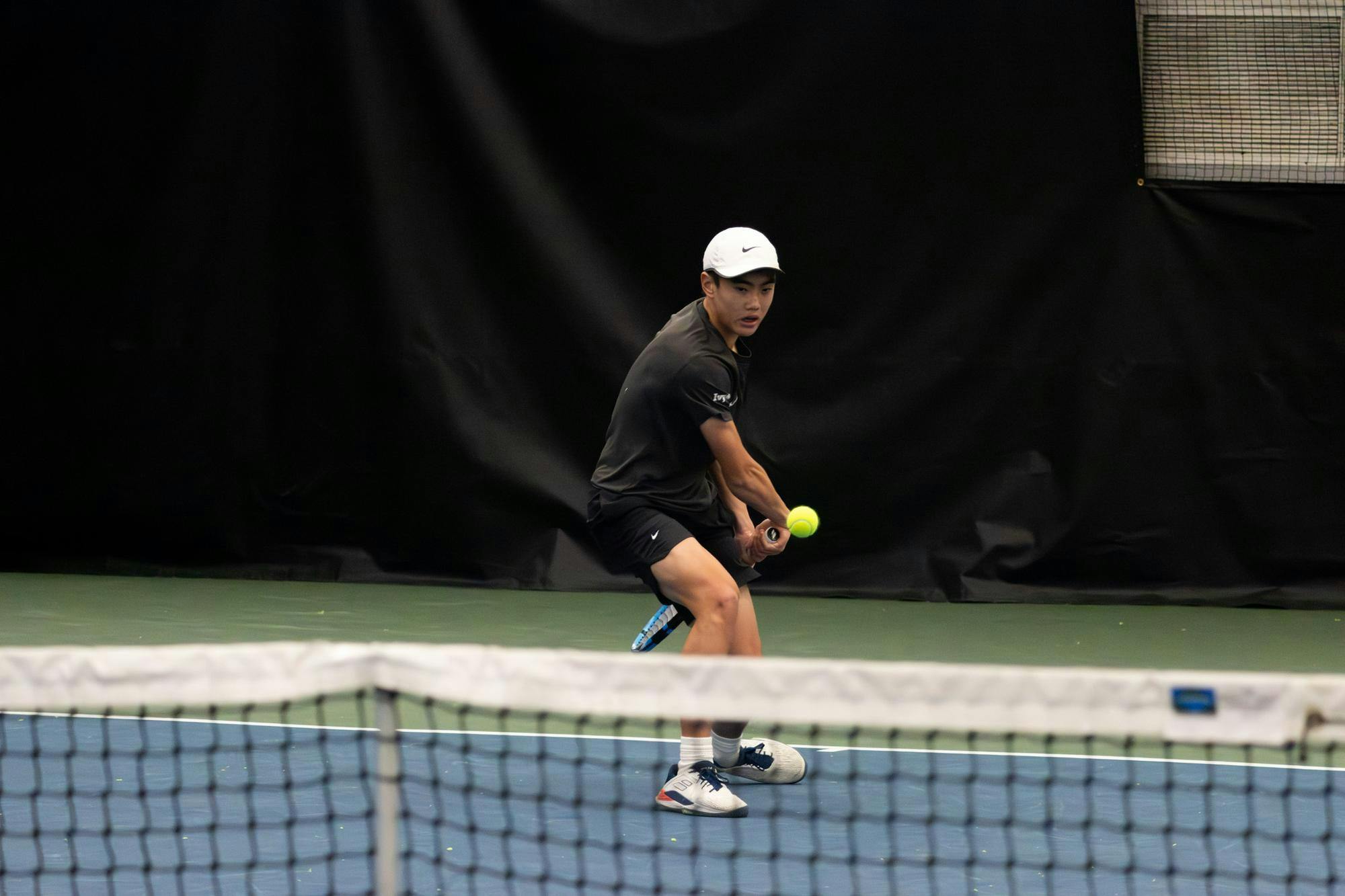 Photo of a Tennis player wearing a black polo shirt and black shorts, getting ready to hit a ball in front of him.

