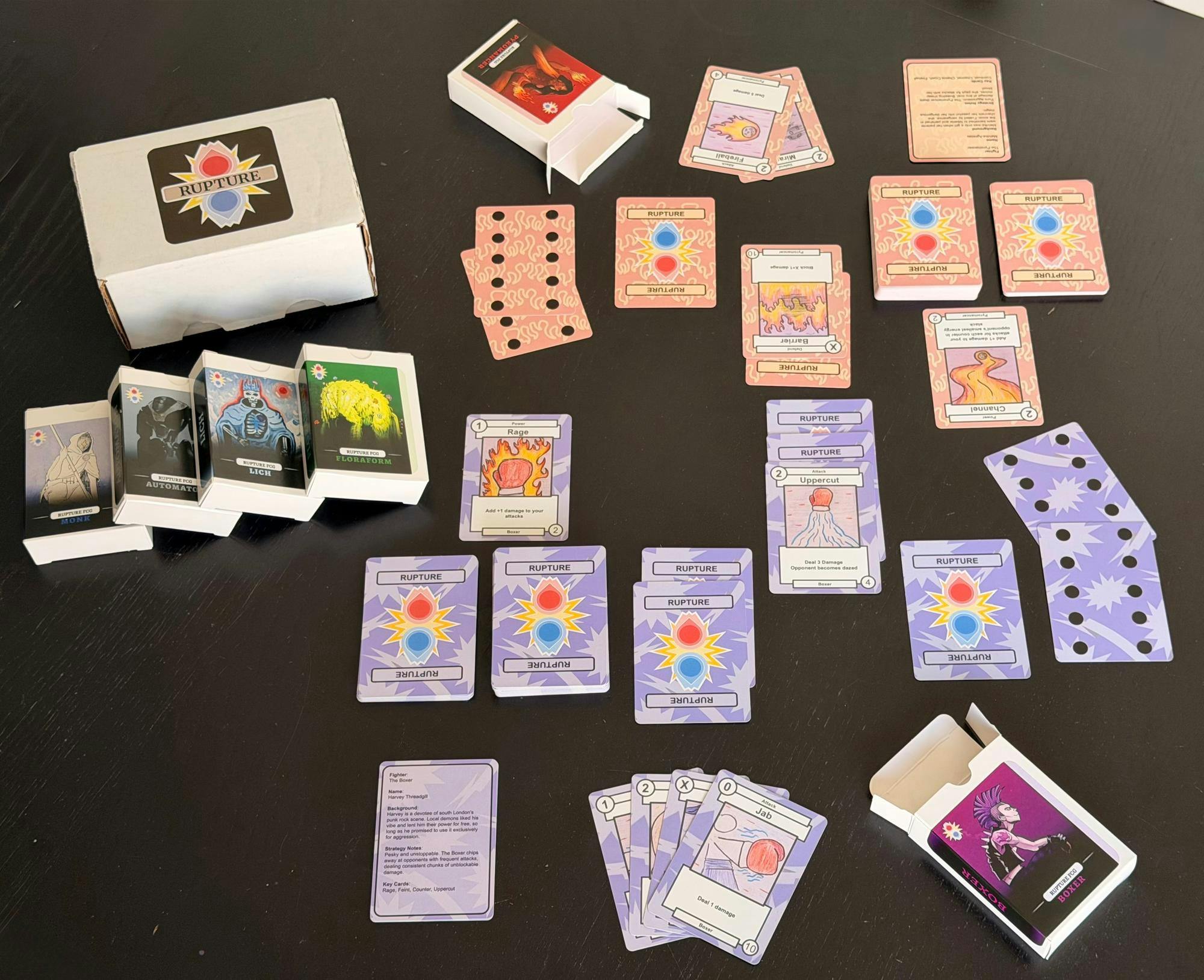 Photo of seven boxes and piles of orange and purple cards, face up and face down. One big box says “Rupture” in all capitals and is surrounded by a red and blue logo. Four boxes are stacked with different characters. Two others are open on different sides of the setup.