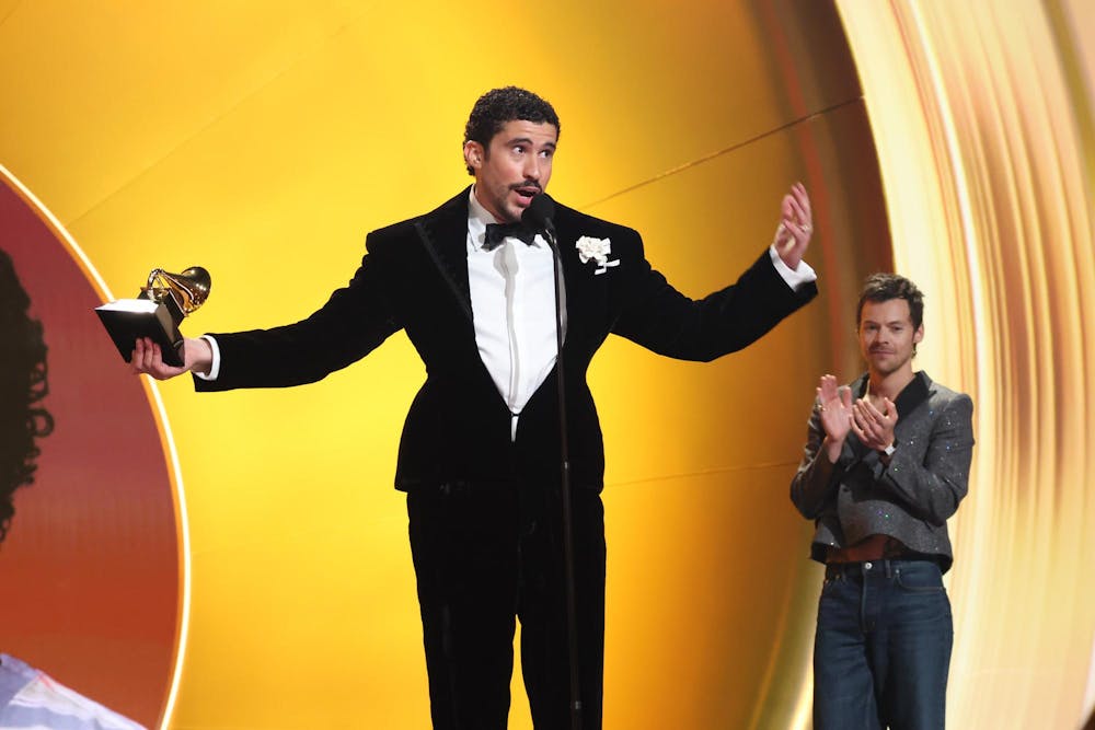 Bad Bunny stands on stage in front of a microphone against a gold backdrop. He is holding a Grammy Award in one hand and wearing a black suit.
