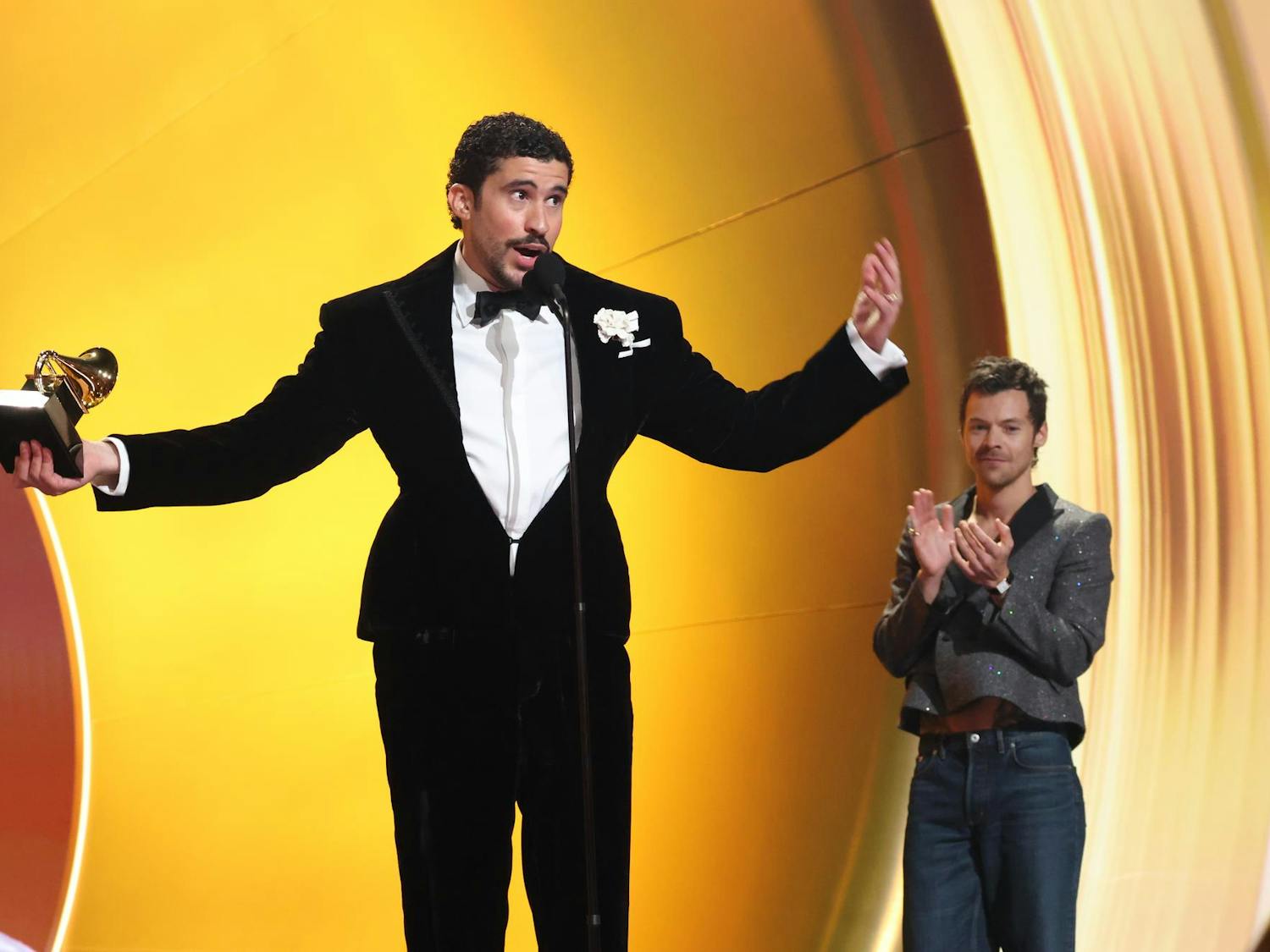 Bad Bunny stands on stage in front of a microphone against a gold backdrop. He is holding a Grammy Award in one hand and wearing a black suit.