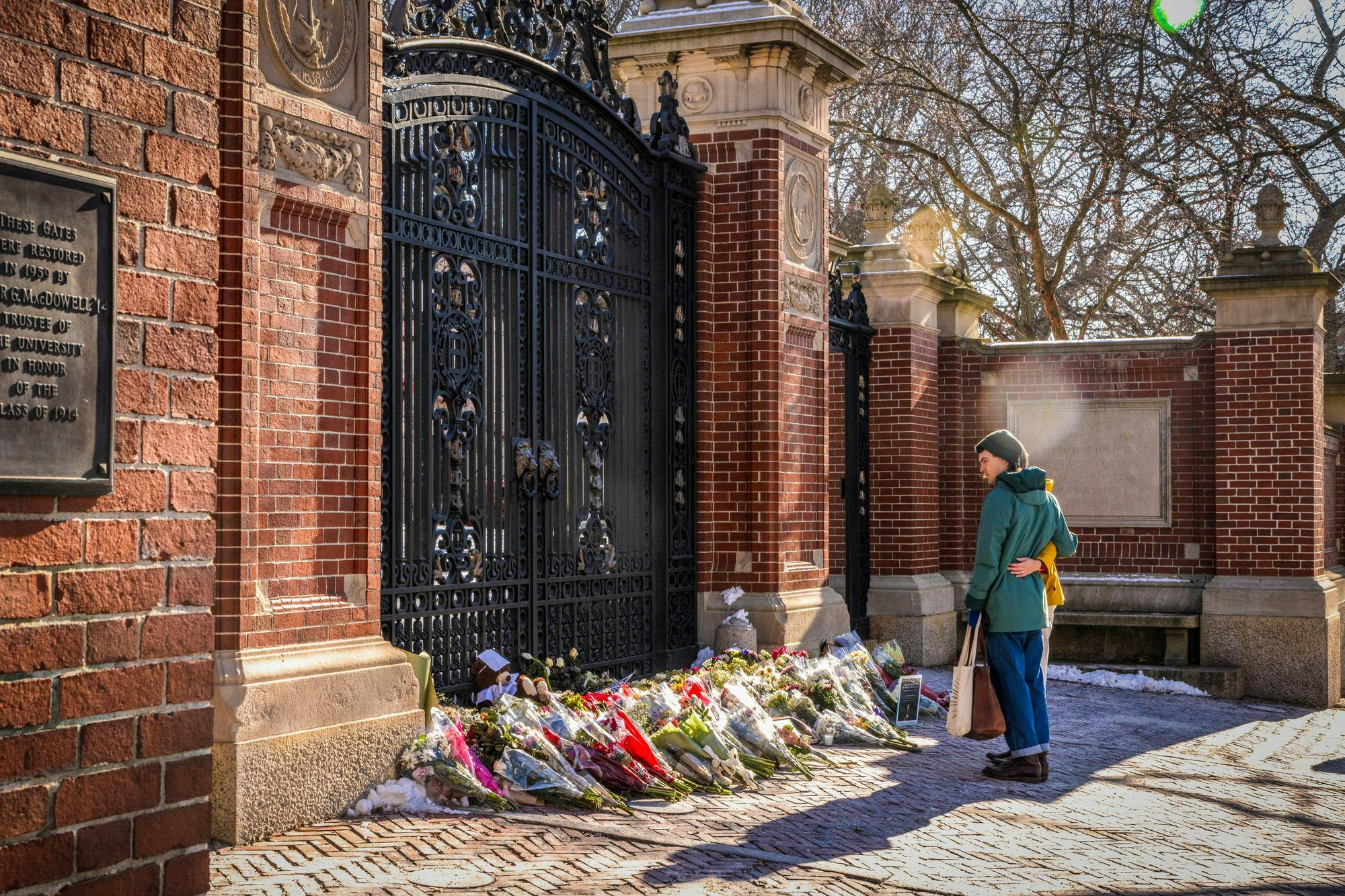 Two people stand together looking down upon a pile of flower bouquets at the foot of Brown University's Van Wickle Gates. 