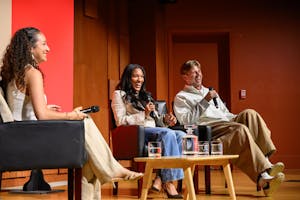 Photo of Tara Davis-Woodhall and Hunter Woodhall laugh holding microphones as they recline in chairs on stage. To the left, Moderator Samantha Chambers '26 smiles and looks towards the audience.
